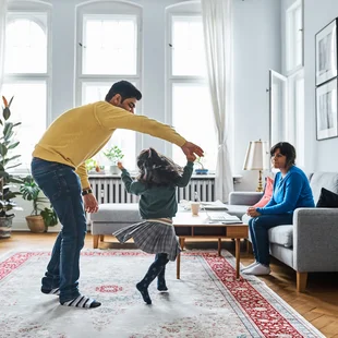 Illustration - Ihr Experte für schönes Wohnen - Mutter schaut Mädchen tanzen mit ihrem Vater auf dem Teppich im Wohnzimmer.